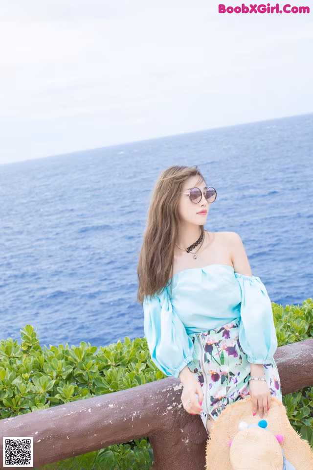 A woman sitting on a railing by the ocean wearing a straw hat.